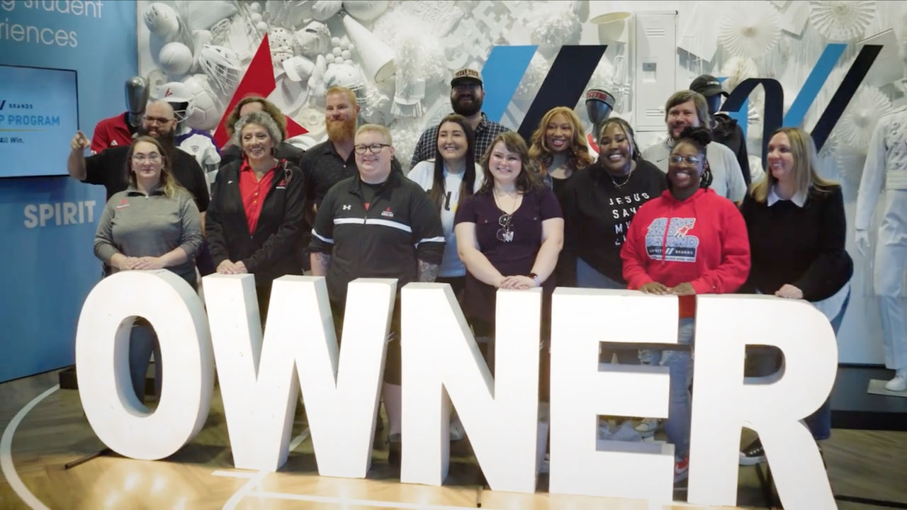 A group of Varsity Brands employees standing behind a large prop that says "OWNER".